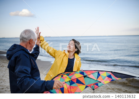 Senior man and his preteen granddaughter preparing kite for flying on sandy beach. Senior man and his preteen granddaughter preparing kite for flying on sandy beach. 140113632