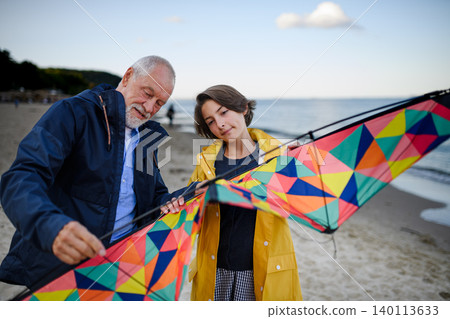 Senior man and his preteen granddaughter preparing kite for flying on sandy beach. 140113633