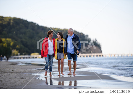 Senior couple holding hands with their preteen granddaughter and walking on sandy beach. 140113639