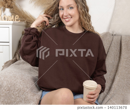 Blank brown oversized sweatshirt mockup on fair-haired young woman sitting on sofa, front view Blank brown oversized sweatshirt mockup on fair-haired young woman sitting on sofa, front view 140113678