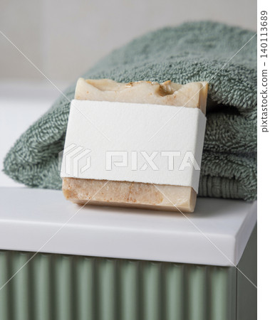 Solid soap with blank paper label mockup near light green towel in bathroom. Unbranded packaging Solid soap with blank paper label mockup near light green towel in bathroom. Unbranded packaging 140113689