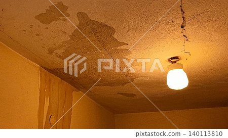 Damaged ceiling with water stains and cracks, illuminated by a single light bulb. A close-up view of a neglected ceiling showing significant water damage, mold growth, and structural cracks, with a Damaged ceiling with water stains and cracks, illuminated by a single light bulb. A close-up view of a neglected ceiling showing significant water damage, mold growth, and structural cracks, with a 140113810