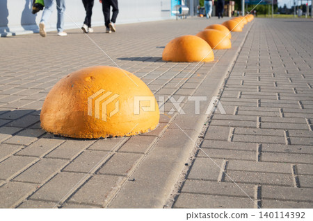 Yellow concrete hemisphere bollards on sidewalk, shopping mall parking lot safety barriers with blurred pedestrians walking Yellow concrete hemisphere bollards on sidewalk, shopping mall parking lot safety barriers with blurred pedestrians walking 140114392