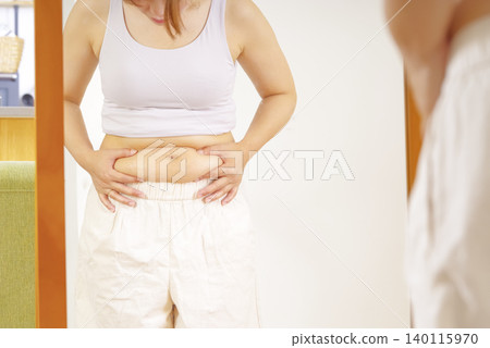 一位女士對著鏡子,擔心自己隆起的肚子。 一位女士對著鏡子,擔心自己隆起的肚子。 140115970