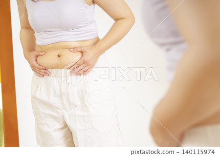 一位女士對著鏡子,擔心自己隆起的肚子。 一位女士對著鏡子,擔心自己隆起的肚子。 140115974