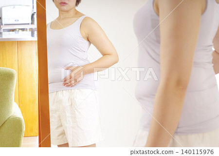一位女士對著鏡子,擔心自己隆起的肚子。 一位女士對著鏡子,擔心自己隆起的肚子。 140115976