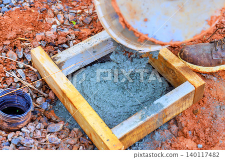 Worker pours concrete from container into wooden mold at construction site during work hours. Worker pours concrete from container into wooden mold at construction site during work hours. 140117482