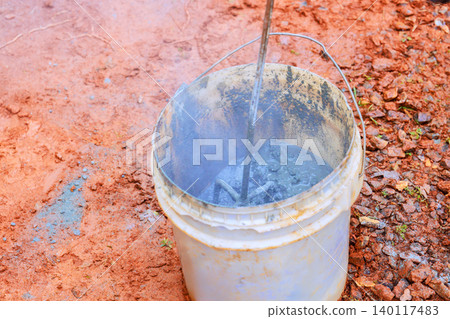 Worker stirs concrete mix in bucket at construction site with tools on dirt surface. Worker stirs concrete mix in bucket at construction site with tools on dirt surface. 140117483