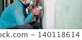 Construction worker uses a handheld power sander to remove old paint dust from an aged wall. The scene highlights surface preparation before repair, repainting, or interior renovation. 140118614