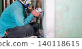 Construction worker uses a handheld power sander to remove old paint dust from an aged wall. The scene highlights surface preparation before repair, repainting, or interior renovation. 140118615
