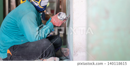 Construction worker uses a handheld power sander to remove old paint dust from an aged wall. The scene highlights surface preparation before repair, repainting, or interior renovation. Construction worker uses a handheld power sander to remove old paint dust from an aged wall. The scene highlights surface preparation before repair, repainting, or interior renovation. 140118615