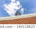 Two pigeons stand close together on the edge of an old tiled roof under a bright blue sky. The scene feels calm, natural, and slightly romantic, with soft clouds in the background. 140118625