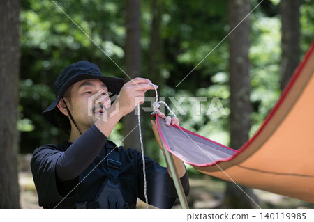 A close-up image of an outdoor nature experience, showing a man enjoying setting up a tarp in the forest. 140119985
