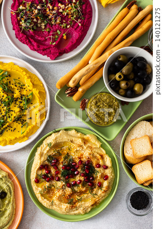 A variety of hummus flavors including turmeric, classic, and broccoli are served with fresh bread, nuts, and garnishes on a colorful charcuterie board, perfect for sharing A variety of hummus flavors including turmeric, classic, and broccoli are served with fresh bread, nuts, and garnishes on a colorful charcuterie board, perfect for sharing 140121275
