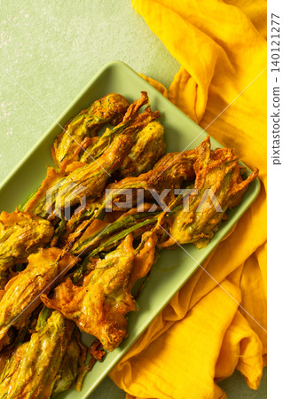 Delicious deep-fried zucchini flowers are neatly arranged on a green platter, showcasing their crispy texture. A bright yellow napkin adds warmth to the scene Delicious deep-fried zucchini flowers are neatly arranged on a green platter, showcasing their crispy texture. A bright yellow napkin adds warmth to the scene 140121277