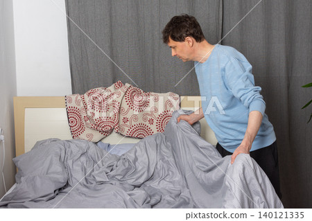 A 53-year-old man is actively changing the bed linen in his bedroom. He is focused on putting fresh sheets on the bed, demonstrating a common home chore A 53-year-old man is actively changing the bed linen in his bedroom. He is focused on putting fresh sheets on the bed, demonstrating a common home chore 140121335