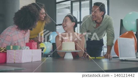 Celebrating team gathering around seated woman with tiara and red dress in office with cake, laptop Celebrating team gathering around seated woman with tiara and red dress in office with cake, laptop 140121442
