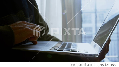 Typing mature woman holding silver laptop at window with sheer curtain, wearing dark long sleeves Typing mature woman holding silver laptop at window with sheer curtain, wearing dark long sleeves 140121490