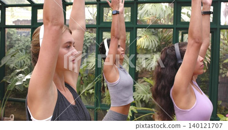 Stretching three women in workout clothes raising arms overhead in greenhouse, with tropical plants Stretching three women in workout clothes raising arms overhead in greenhouse, with tropical plants 140121707