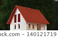 Showing white scale model house sitting on flat table near blurred green plants, featuring red roof 140121719