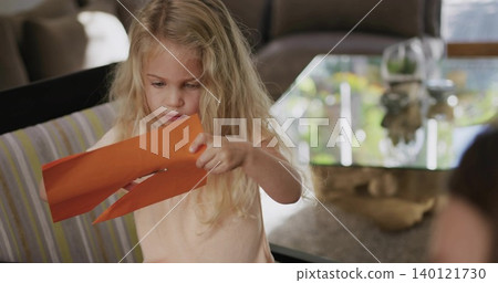 Holding orange craft paper, blonde child studying cut strips at home, wearing peach sleeveless top 140121730