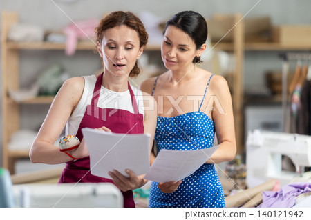 Female tailor shows paper sketch of dress to woman client Female tailor shows paper sketch of dress to woman client 140121942