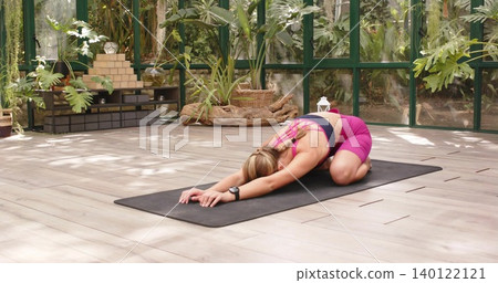 Stretching woman wearing pink sportswear folding forward on black mat in greenhouse with wristwatch 140122121