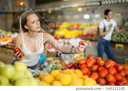 Woman carefully selects fresh tomatoes, standing near the counter in supermarket Woman carefully selects fresh tomatoes, standing near the counter in supermarket 140122185