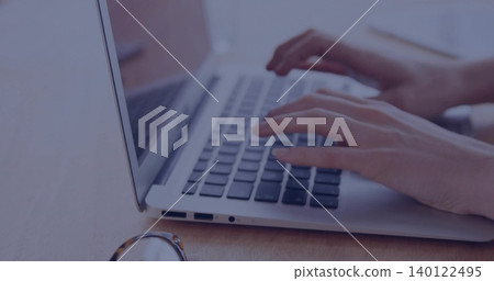Typing woman's hands pressing keys on laptop keyboard at wooden desk, with eyeglasses nearby 140122495