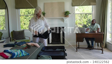 Packing senior woman in white blouse folding clothes into open suitcase in bedroom, laptop nearby Packing senior woman in white blouse folding clothes into open suitcase in bedroom, laptop nearby 140122562