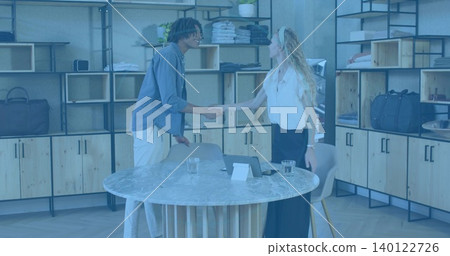 Shaking hands two staff in business attire leaning across marble table in showroom, tablet visible Shaking hands two staff in business attire leaning across marble table in showroom, tablet visible 140122726