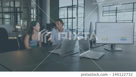 Talking pair wearing sleeveless top and blue shirt at table in office with silver laptops, monitor Talking pair wearing sleeveless top and blue shirt at table in office with silver laptops, monitor 140122732