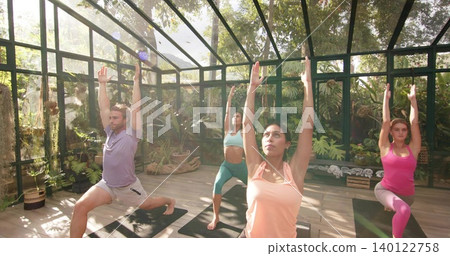 Practicing four adults in activewear holding lunge poses in glass greenhouse with mats, plants Practicing four adults in activewear holding lunge poses in glass greenhouse with mats, plants 140122758
