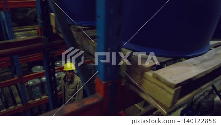 Inspecting worker wearing yellow hard hat, quilted vest looking up in storage aisle at blue barrels Inspecting worker wearing yellow hard hat, quilted vest looking up in storage aisle at blue barrels 140122858