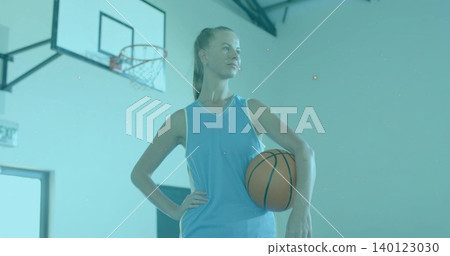 Holding ball at hip, female basketball player wearing light blue uniform posing in gym with hoop Holding ball at hip, female basketball player wearing light blue uniform posing in gym with hoop 140123030