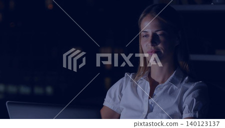 Working mid-adult woman leaning toward open laptop at night desk, wearing white blouse, copy space 140123137