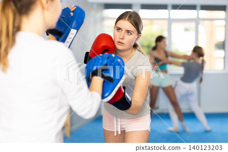 Adolescent girl in boxing gloves punching pads in hands of coach in gym Adolescent girl in boxing gloves punching pads in hands of coach in gym 140123203