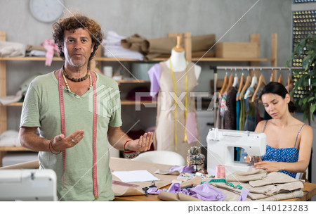 Middle-aged male dressmaker inviting clients while girl sew Middle-aged male dressmaker inviting clients while girl sew 140123283