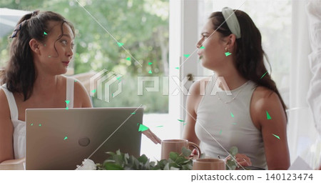 Discussing women in tank tops, headband, layered necklaces at table by glass door with laptop, mugs 140123474