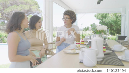 Sitting three women chatting and sipping tea on covered patio by backyard pool, holding smartphone Sitting three women chatting and sipping tea on covered patio by backyard pool, holding smartphone 140123494