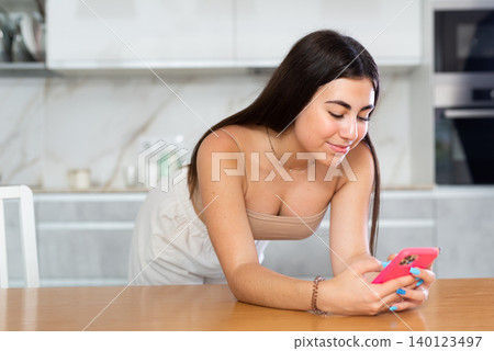 Young woman leaning on kitchen table and looking at her phone Young woman leaning on kitchen table and looking at her phone 140123497