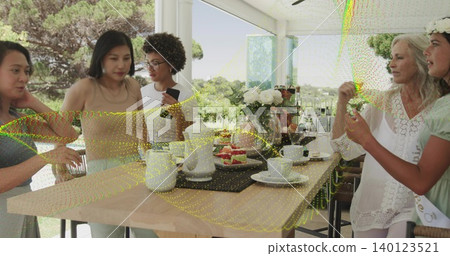Gathering five adults wearing light green dress and white blouse at sunroom table, with teapots 140123521