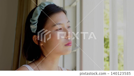 Gazing Korean woman by window at home, wearing pale tank top, pale-green padded headband, necklace 140123585