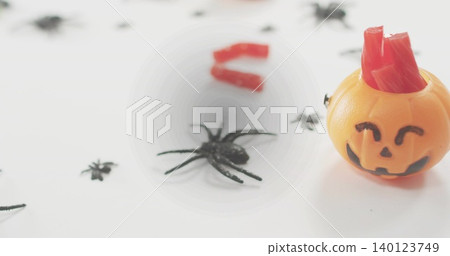 Sitting jack-o'-lantern on white table, holding red gummy strips with black spiders, copy space Sitting jack-o'-lantern on white table, holding red gummy strips with black spiders, copy space 140123749