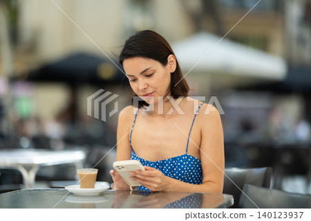 Brunette texting on smartphone screen during breakfast on cafe terrace. Drinking coffee Brunette texting on smartphone screen during breakfast on cafe terrace. Drinking coffee 140123937