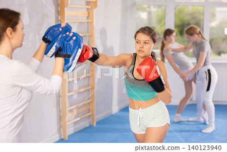 Focused teenage girl in boxing gloves practicing punches on mitts with coach Focused teenage girl in boxing gloves practicing punches on mitts with coach 140123940