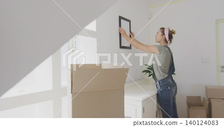 Hanging midadult woman in overalls placing frame above cabinet at home with boxes, copy space Hanging midadult woman in overalls placing frame above cabinet at home with boxes, copy space 140124083