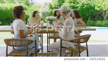 Chatting group of five women sitting around rectangular wooden table on patio by pool, with roses Chatting group of five women sitting around rectangular wooden table on patio by pool, with roses 140124201