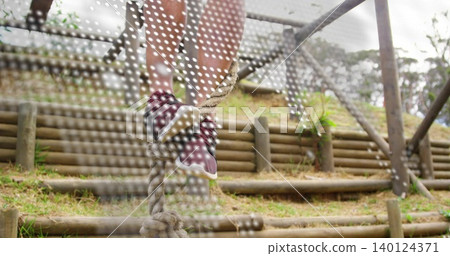 Climbing legs of man ascending knotted rope at wooded obstacle park, maroon shoes shorts white mesh 140124371