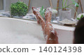 Reclining woman extending bare legs over bathtub rim at window, with potted plants, foam, tray 140124398